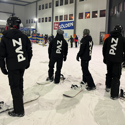 PAZ-Team in Schneeanzügen vor Förderband in Skihalle, Rückenansicht