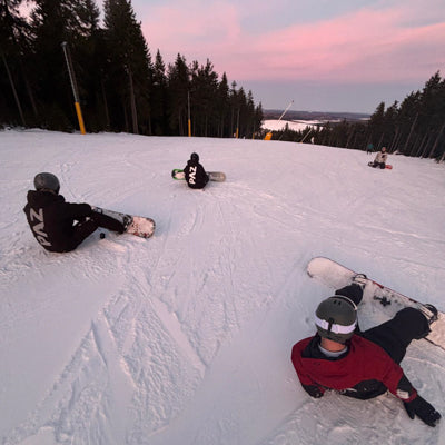 Zwei Snowboarder in PAZ Schneeanzügen auf Piste bei Sonnenuntergang