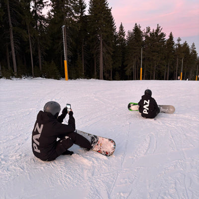 Zwei Snowboarder im PAZ Schneeanzug sitzend auf der Piste, Blick Richtung Wald